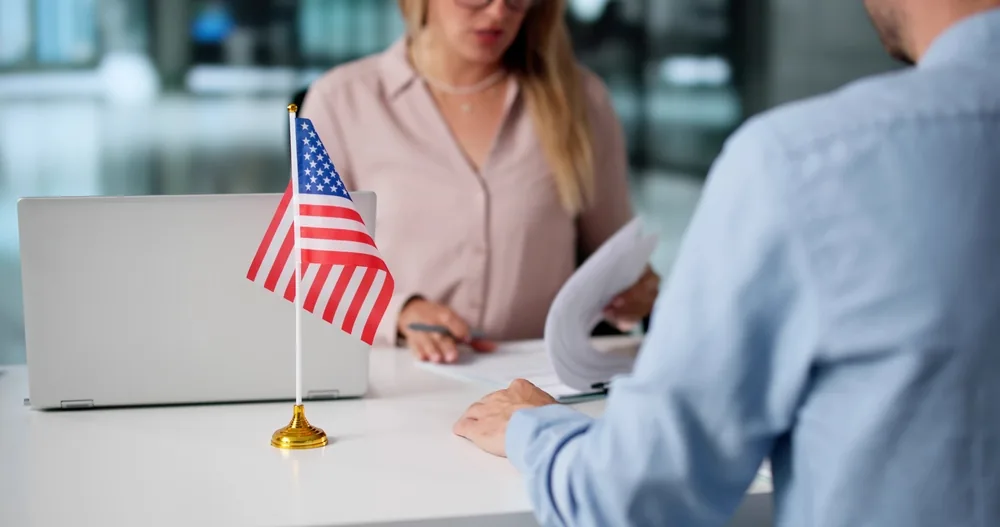 An applicant hands documents across a desk with a small U.S. flag, reviewing green card renewal paperwork with an immigration professional