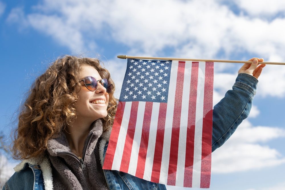 A green card applicant smiling as she holds a small United States flag against a blue sky