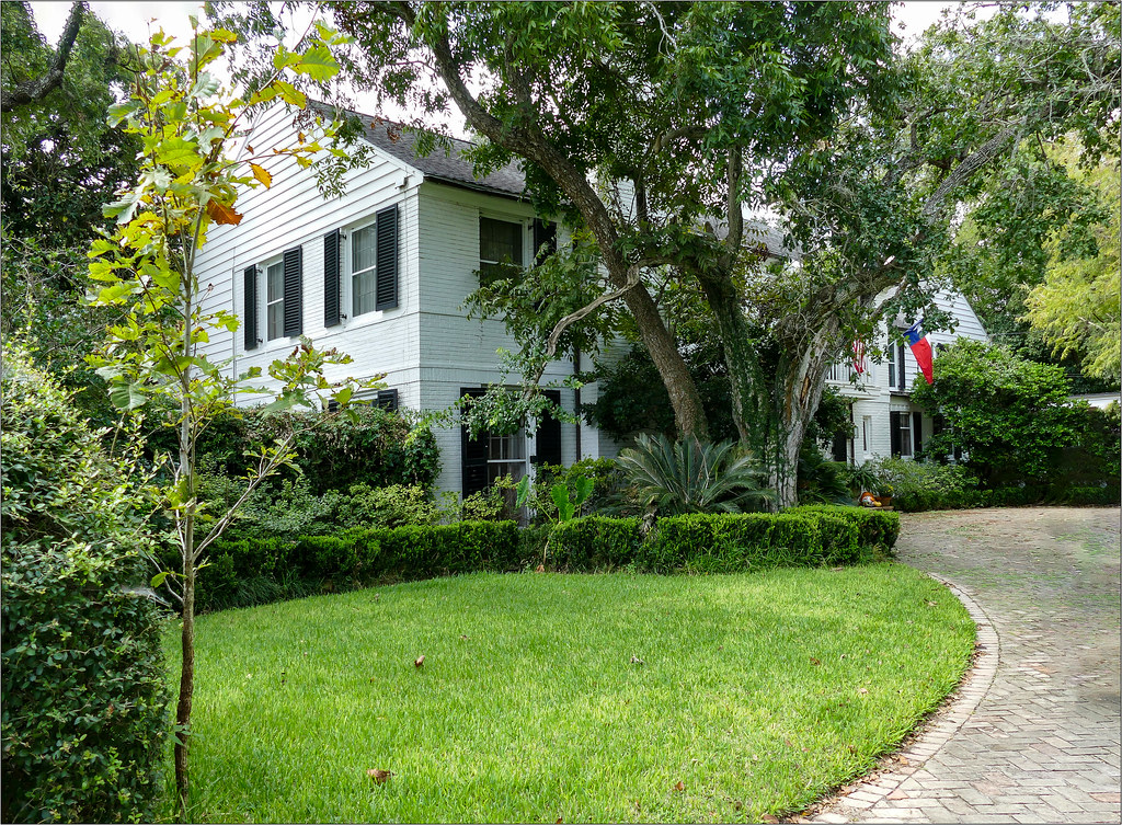 A white clapboard house behind a lawn and mature trees in Victoria, Texas.