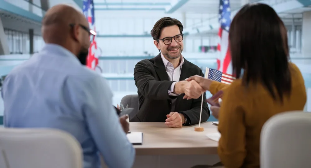 A couple sits across a desk from a smiling official in glasses, who shakes the woman's hand beside a small U.S. flag